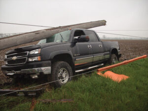 A shattered utility pole remains atop the damaged pickup truck with de-energized lines still attached.
