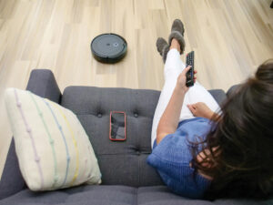 A woman sits on a couch while a robotic vacuum moves on the floor in front of her.