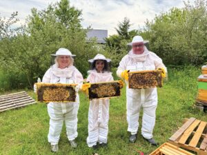 Three people in white bee keeping suits and netted hats smile while holding honey supers - rectangles filled with honey comb and bees.