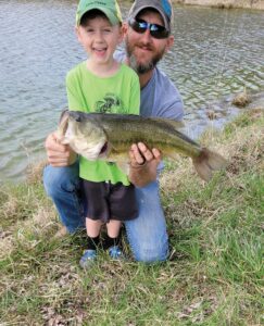 A father knees behind his son, both smiling while holding a fish the boy caught from the lake behind them.