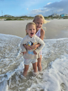 two small blond girls, presumably sisters, stand on the beach, their feet covered by a small wave of water. The older girl is behind the smaller, and has her arms around her. They're both smiling with delight of the water rushing over their feet.