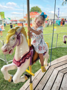 A blond girl with a blue bow in her hair smiles from where she is perched upon a carousel horse.