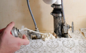 A hand holds a can of spray foam, sealing the space between plumbing and the bottom of a vanity