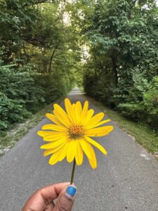 A woman's hand holds a yellow flower before a narrow road. Chipped blue nail polish adorns her thumbnail