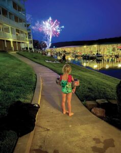 A small blond girl wearing water wings and a green swimsuit stands with her back to the camera as she stares over a marina at fireworks
