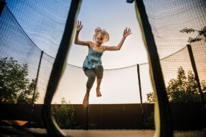 Child girl jumping high on big trampoline outside in garden on summer sunset.