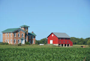 Across a green field a brick Italionate home stands beside a bright red barn