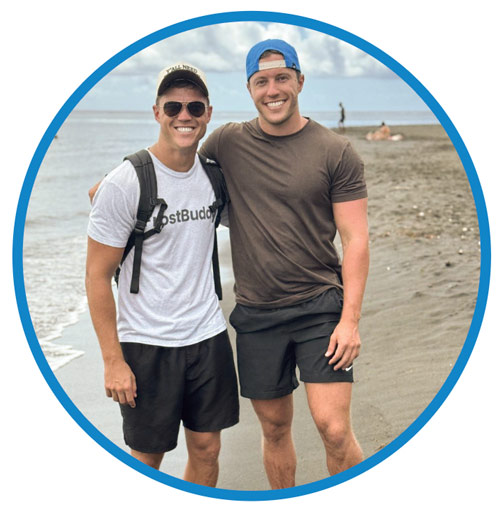 Two smiling young men pose on a beach.