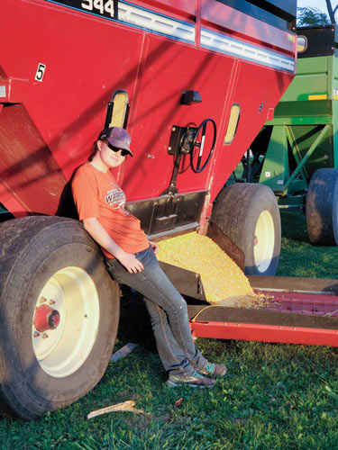 A young woman in a hat, sunglasses, t-shirt and jeans leans against a harvester.
