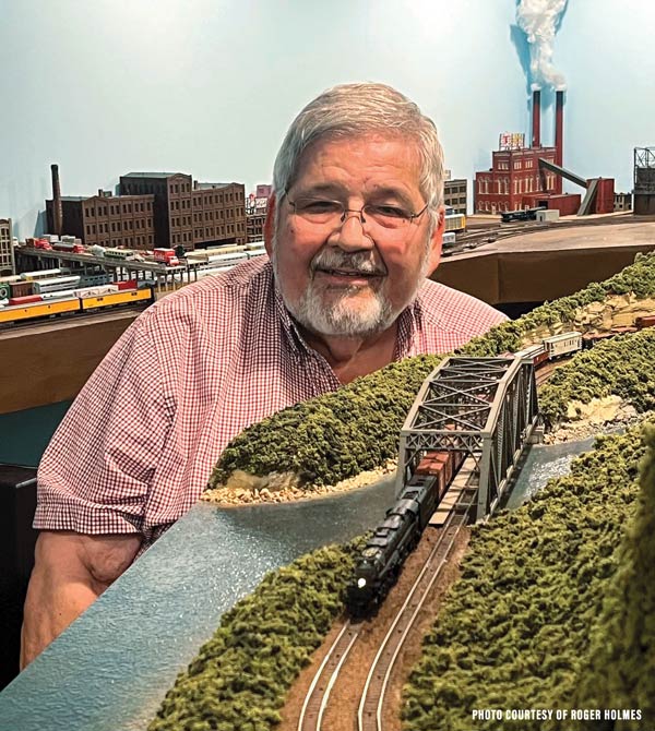 A smiling, bearded man poses by a model train landscape he created.
