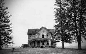 Black and white photo of a dilapidated two-story farmhouse framed by pine trees.