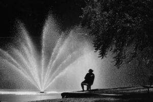 The black and white photo captures, from a distance, a person sitting in a bench before a fountain