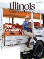A woman in a cowboy hat and boots sits on the edge of a white converted school bus where behind her a ton of orange pumpkins are stacked