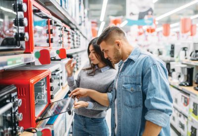 A young couple stand in a store aisle and look at a bright red toaster oven.