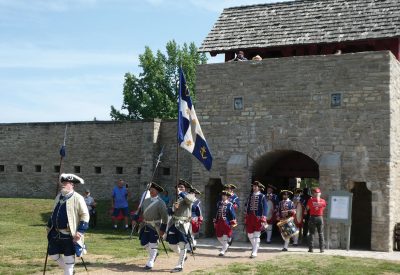 A line of men in French military costumes and flags march out of the fort's gate