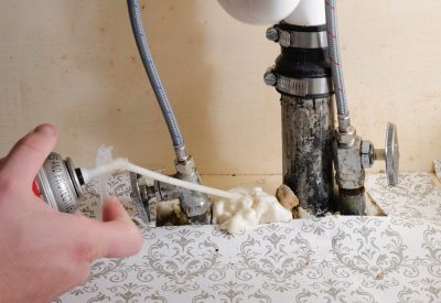A hand holds a can of spray foam, sealing the space between plumbing and the bottom of a vanity