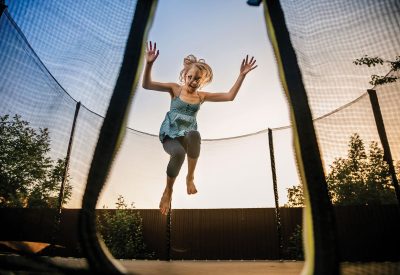 Child girl jumping high on big trampoline outside in garden on summer sunset.