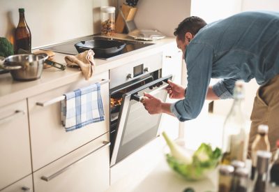 Portrait of gentleman in denim shirt closing door of oven while looking at frying pan with beef steak